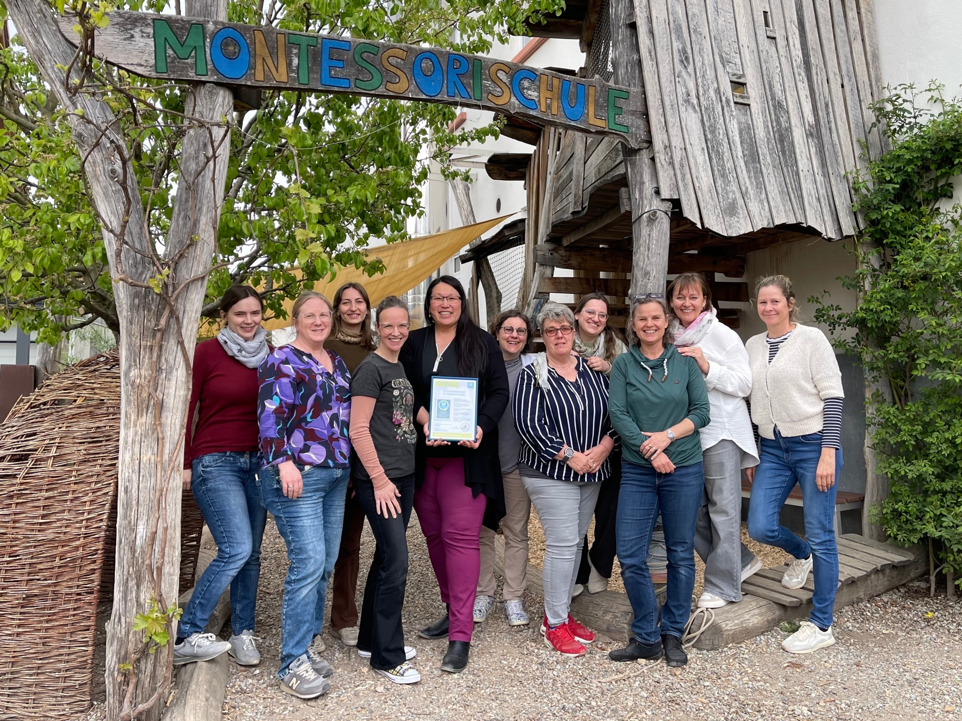 Das Team der Montessori Schule Worms steht gemeinsam unter einem Holzschild und präsentiert die QR-Anerkennungsurkunde