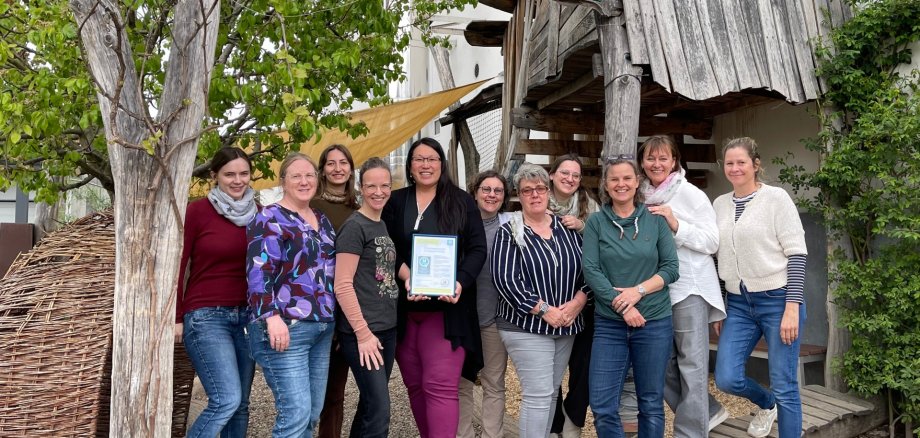 Das Team der Montessori Schule Worms steht gemeinsam unter einem Holzschild und präsentiert die QR-Anerkennungsurkunde